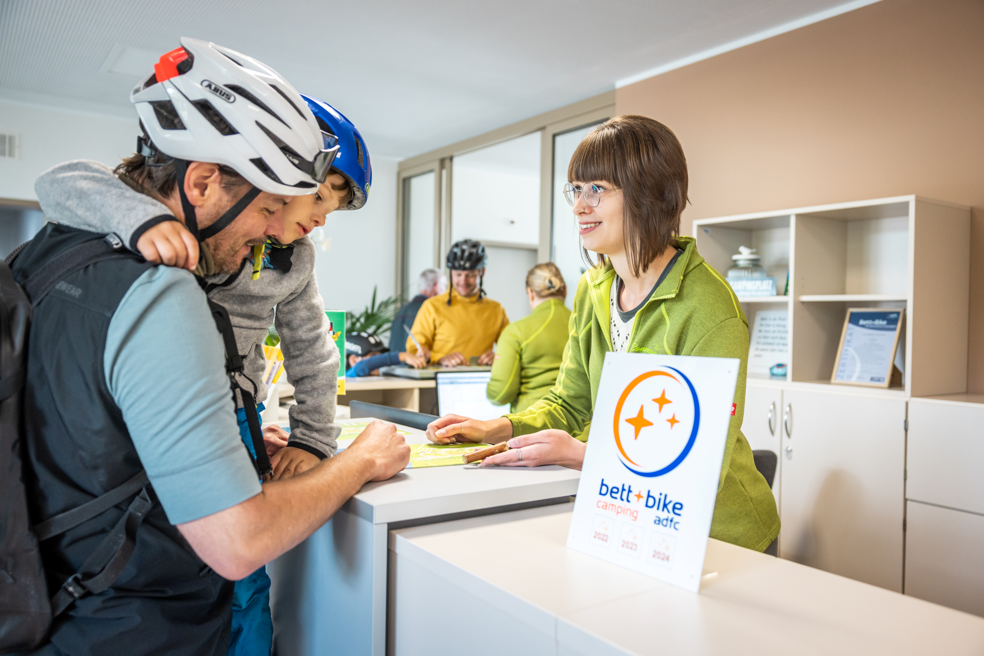 Bett+Bike - Rezeption Camping Ein Vater mit Kind auf dem Arm informiert sich an einer Rezeption eines Campingplatzes, beide tragen Fahrradhelme. Im Vordergrund steht ein Bett+Bike-Camping-Schild.
